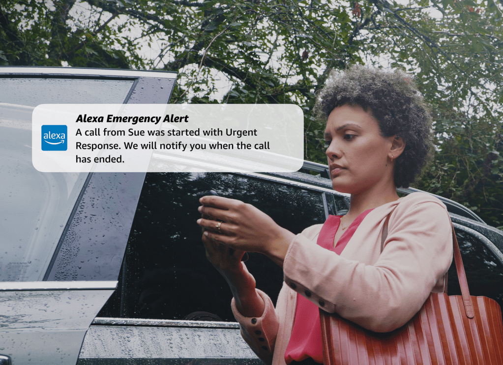 A woman is standing by her car and looking at her smart phone. A text bubble is overlaid the image and says, "Alexa Emergency Alert: A call from Sue was started with Urgent Response. We will notify you when the call has ended."